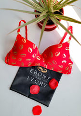 overhead shot of red and gold yemaya cowrie shell print bra laying on satin gift bag with ebony and ivory logo and rose petals on white background with plant in red pot placed above